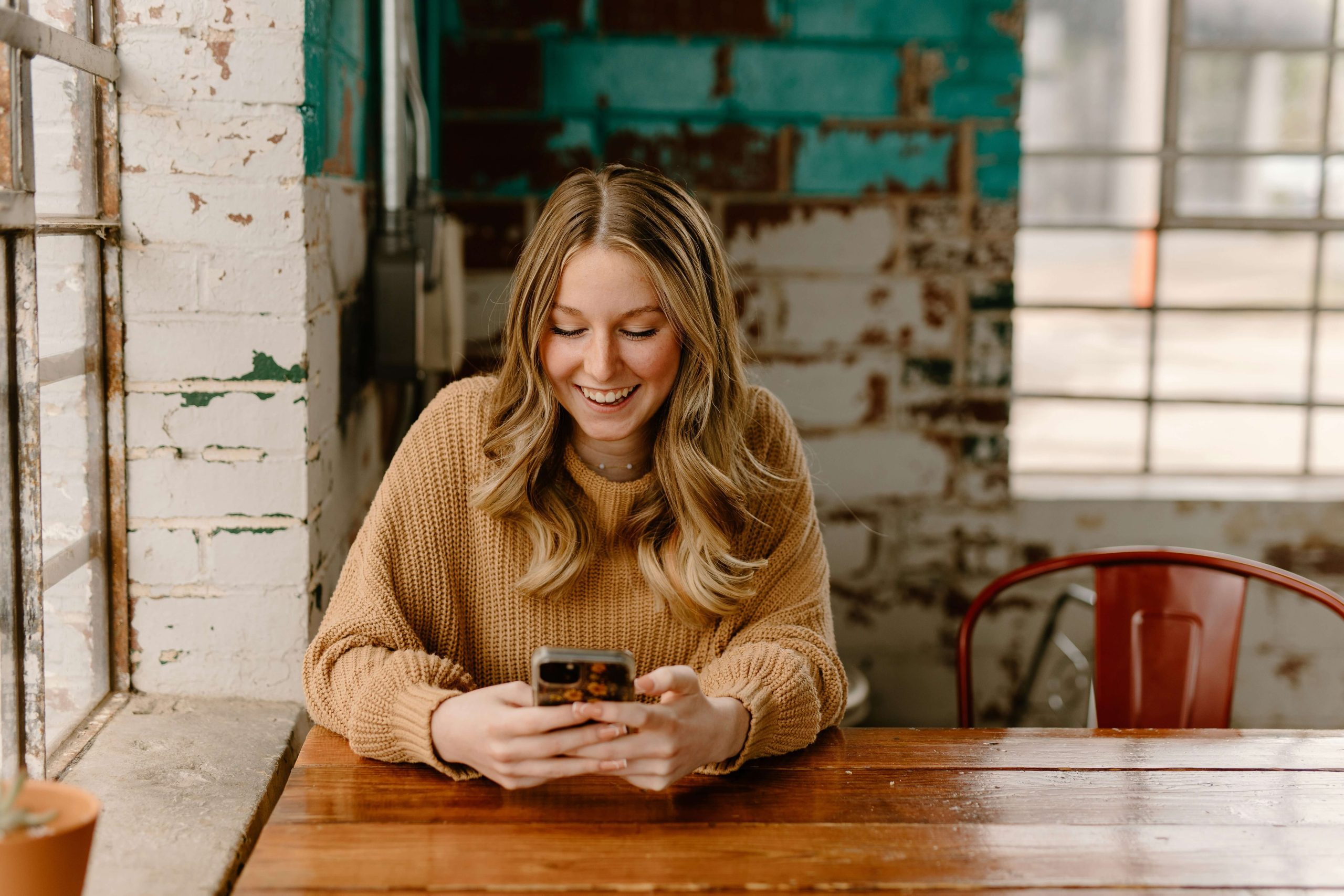 A young woman sitting in a café, holding a phone with both hands and smiling at the screen. A young woman sitting in a café, holding a phone with both hands and smiling at the screen.