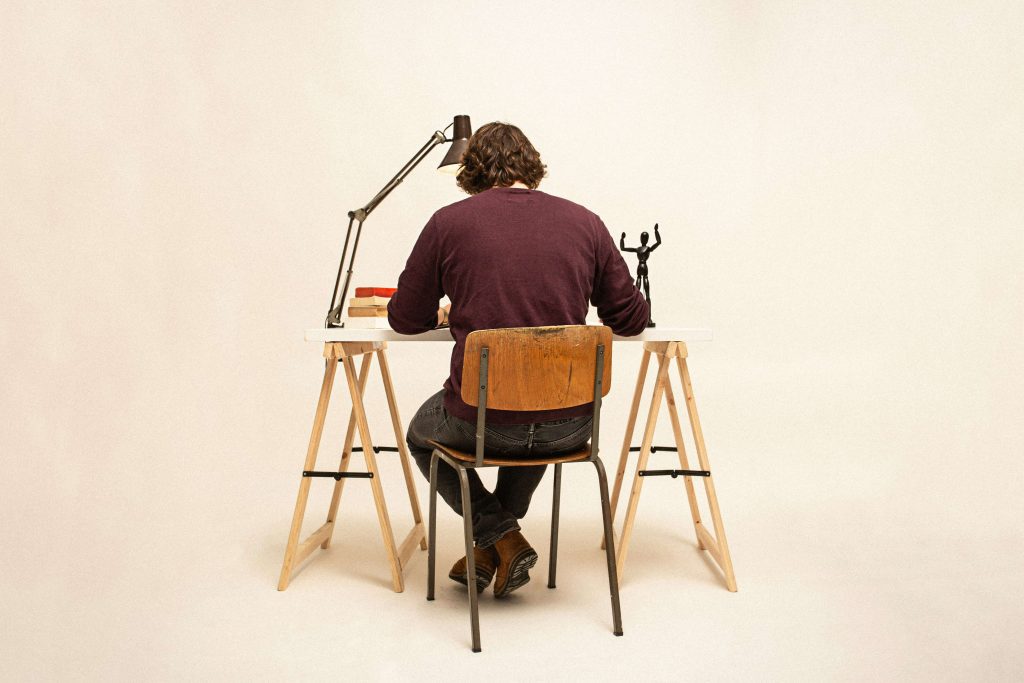 A man in a brown jumper sitting on a wooden chair, working at a desk.
