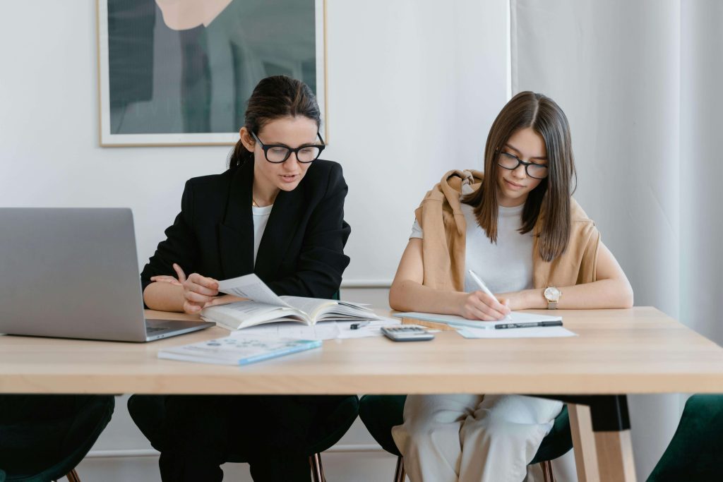 Two women sitting side by side with a table in front of them. One is writing, another is looking at a book in front of her. Both focused on learning.
