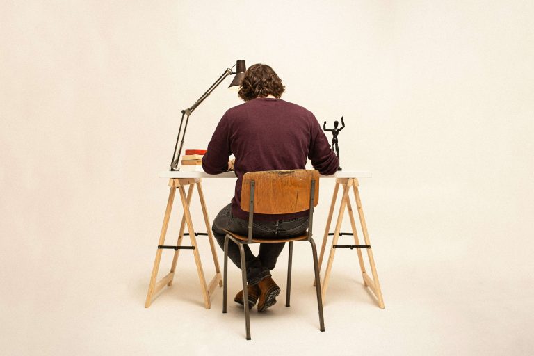 A man in a brown jumper sitting on a wooden chair, working at a desk.