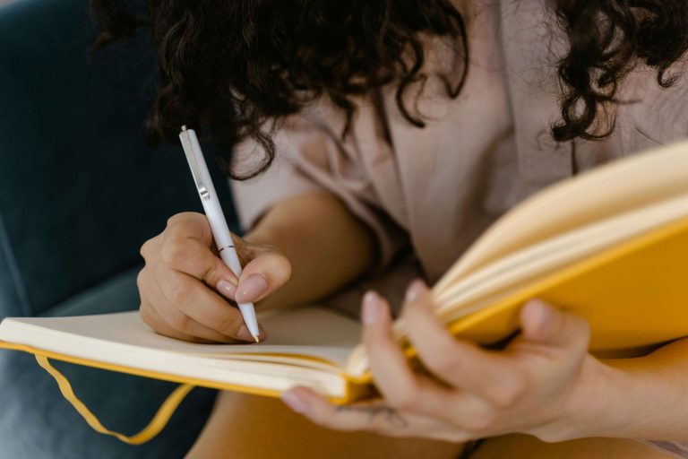 A woman writing on a mustard yellow notebook.