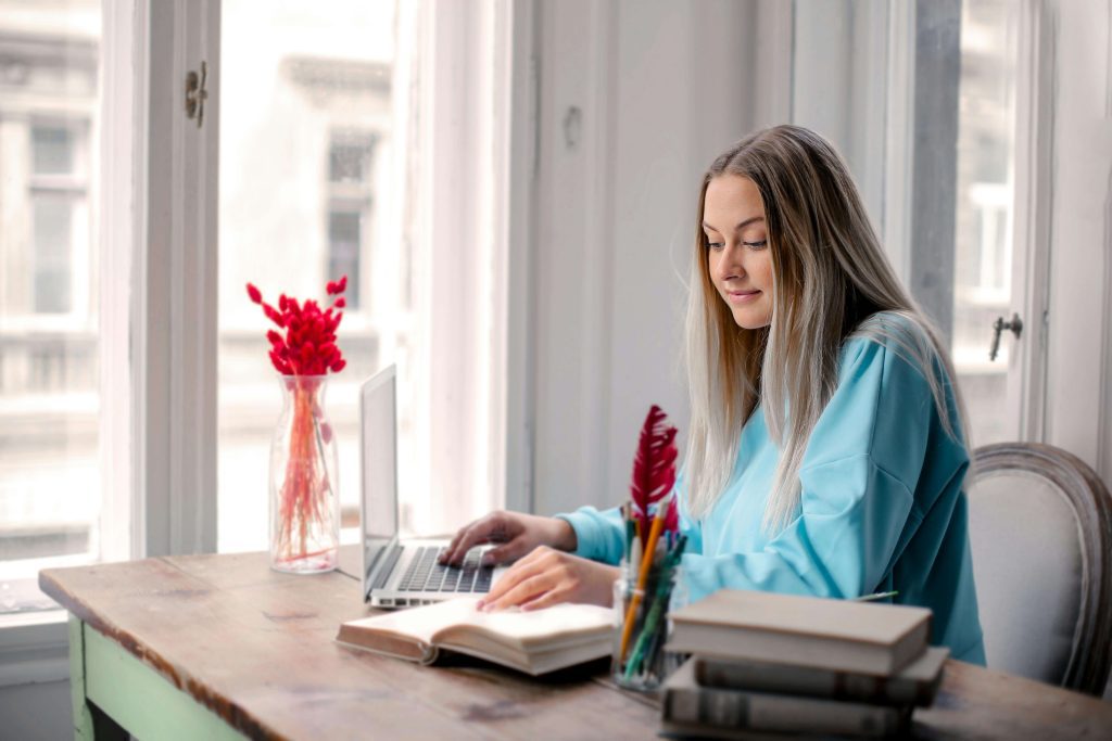 A writer with one hand placed on a laptop in front of her, and the other in an open book.