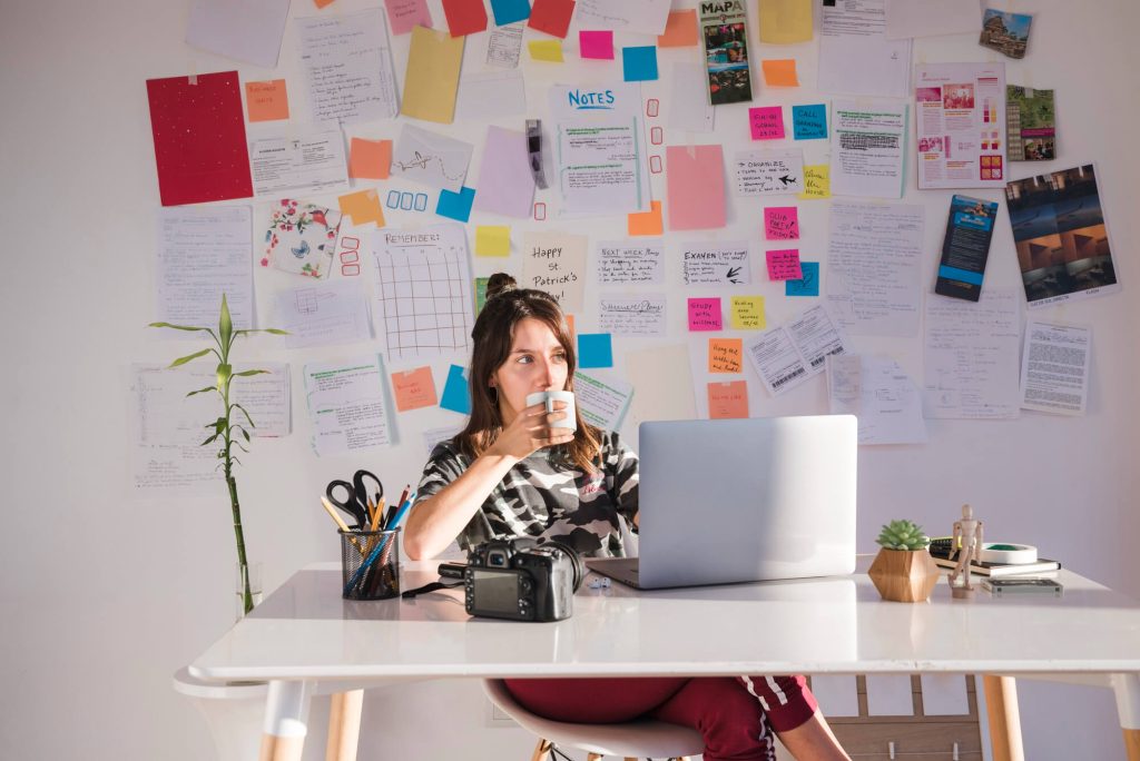 A woman drinking a cup of coffee, sitting behind a chaotic display of post-its and notes.