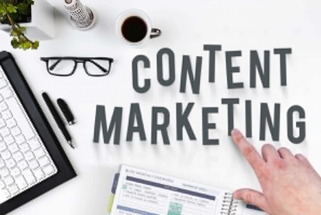 A finger pointing at letters on the table stating CONTENT MARKETING. Scattered around are a coffee mug, glasses, pen, keyboard, notebook with headline BLOG MONTHLY OVERVIEW, a candle, and a green leafy potplant
