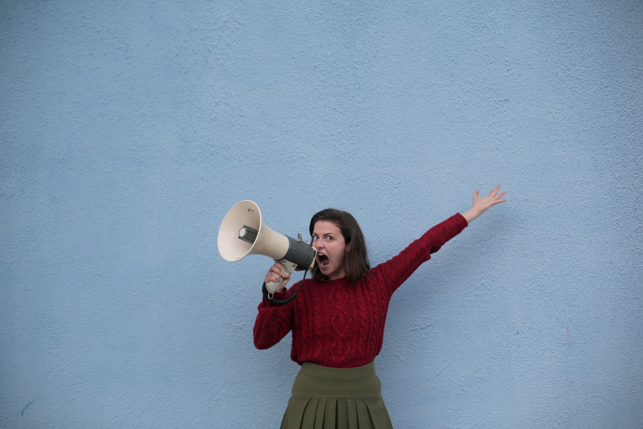 Woman shouting into her megaphone, looking at the camera in front of a muddy-blue wall, her left hand raised above her head. She's wearing a green skirt and a red jumper and she has a very intense look on her face, with her brown hair cut at her shoulders. This image was repurposed to represent using the correct voice and tone.