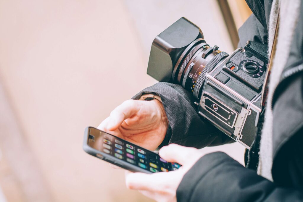 A close-up of a person holding a vintage camera with a large lens and an attached digital viewfinder, as well as a smartphone in their other hand.