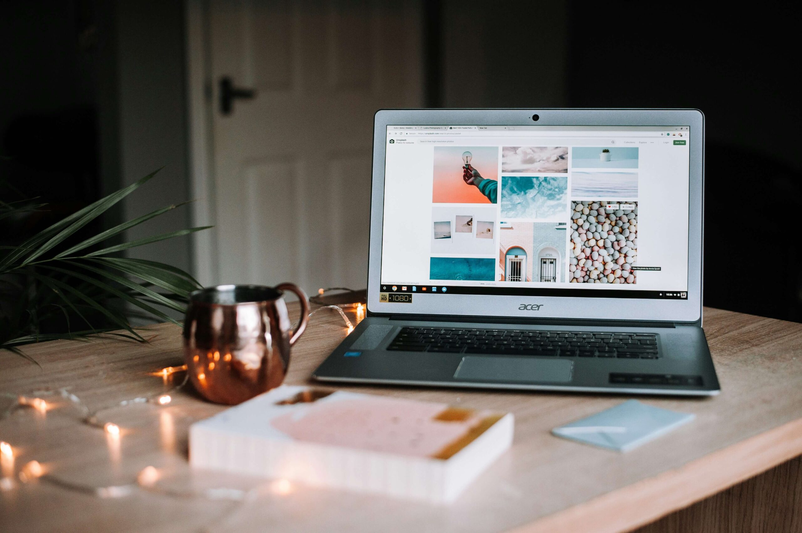 Laptop on a desk with the screen open to a blog displaying different areas of the world. There is a book in front of the laptop with a candle and string of lights on the desk.