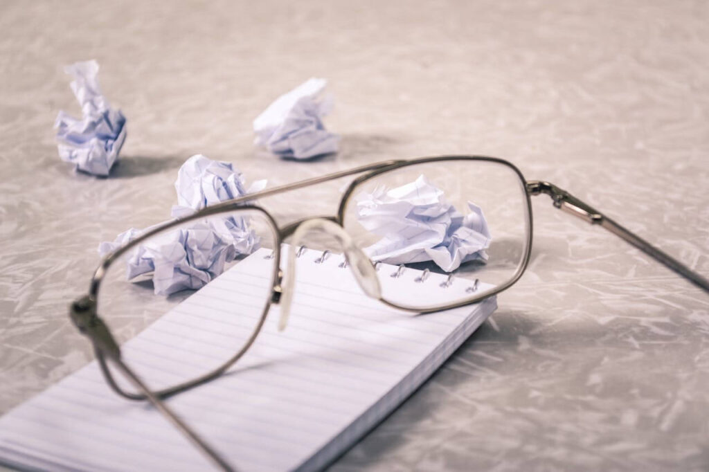 A pair of glasses on a table on top of a notepad. A few scrunched up pages lie in the background showing the frustrations some people go through during content ideation.