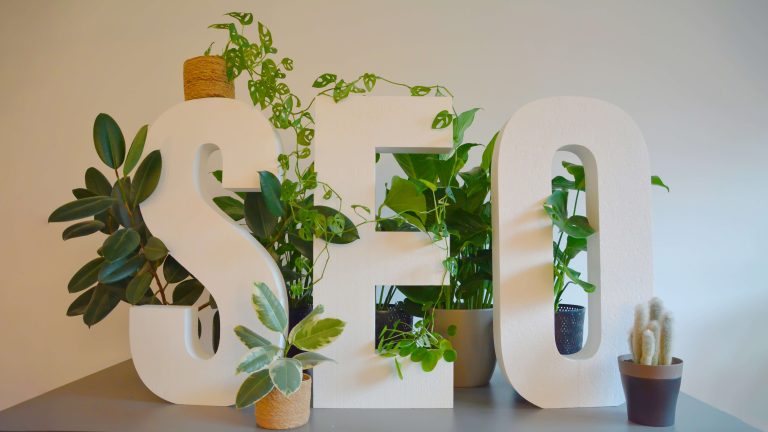 The words "SEO" are prominently displayed on a gray desk. The letters are adorned with an abundance of lush green plants, creating a refreshing and natural atmosphere.