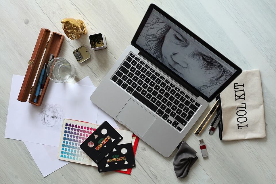 Cluttered table of a graphic designer with a pen, a sharpener, a laptop with a drawing on the screen, a sketch, color swatches, and a pencil case saying toolkit